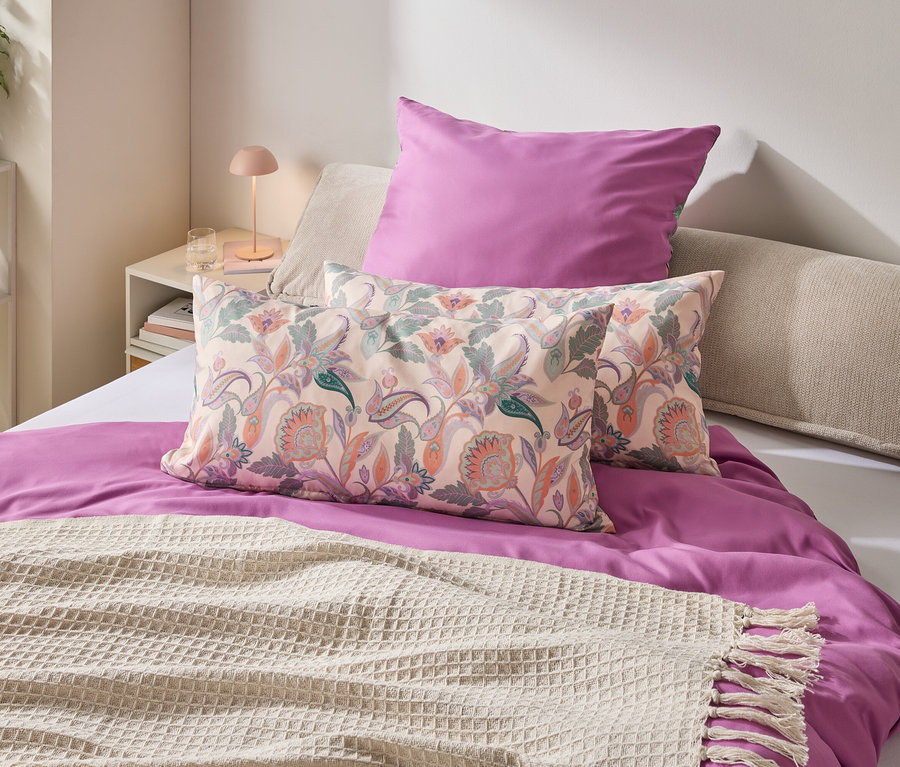 Une chambre avec un lit orné d'une housse de couette violette, d'oreillers à motifs floraux et d'une couverture crème à franges.