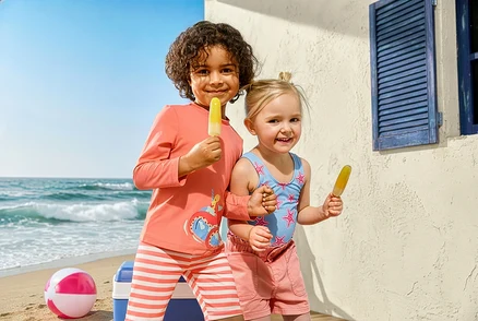 Deux enfants avec des glaces à la plage. La fille porte un maillot de bain pour enfant, étoiles de mer et un short en jean pour enfant. Le garçon porte un ensemble pour enfant avec indice de protection UV 50+, poulpe.