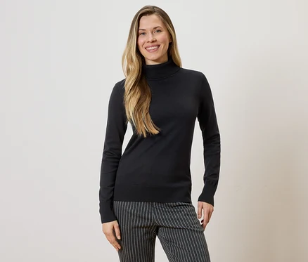 Une femme aux longs cheveux blonds portant un pull à col roulé noir et un pantalon rayé sur un fond clair.