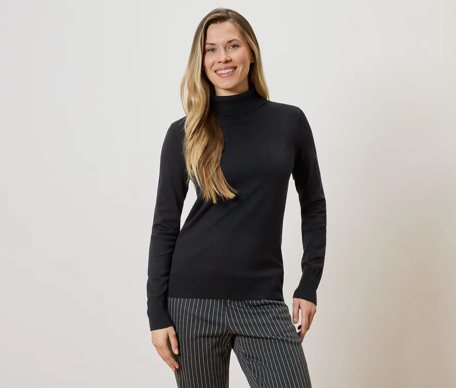 Une femme aux longs cheveux blonds portant un pull à col roulé noir et un pantalon rayé sur un fond clair.