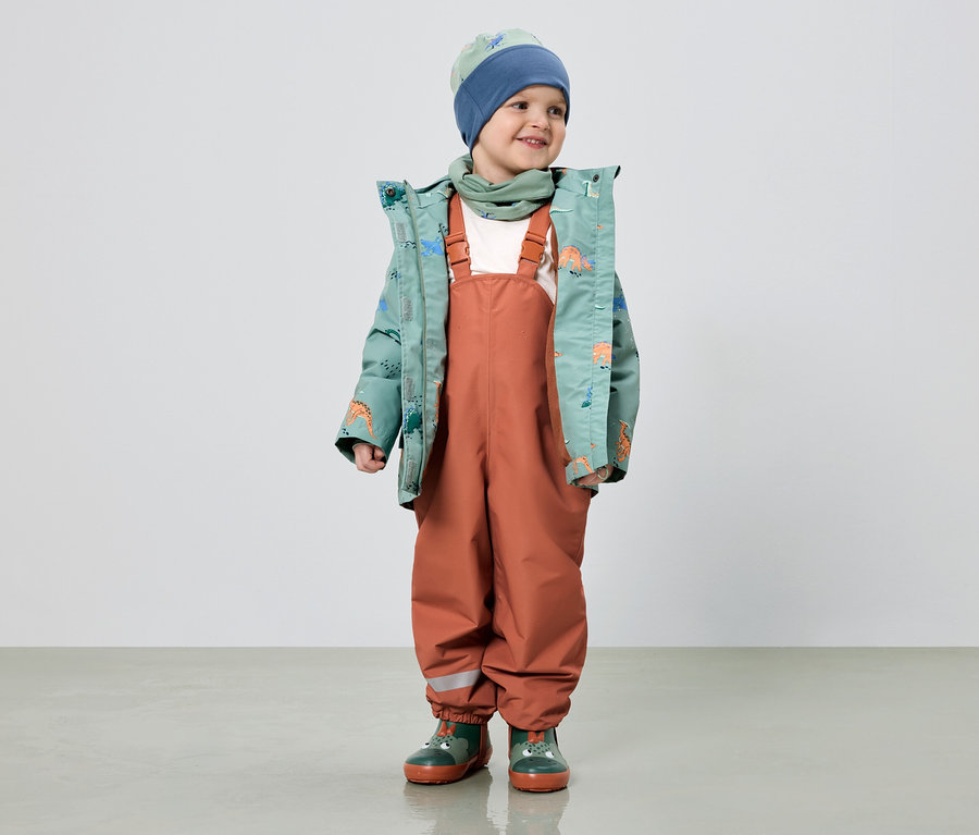 Un garçon souriant porte une veste verte à motifs de dinosaures, une salopette orange, un bonnet bleu et des bottes en caoutchouc vertes.