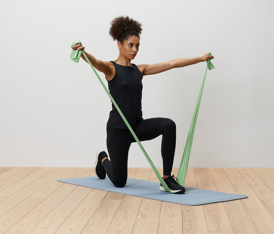 Une femme agenouillée sur un tapis de yoga bleu étire une bande de résistance verte.