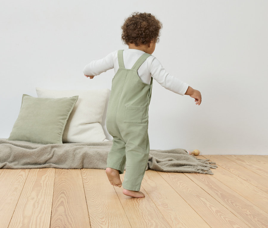 Un jeune enfant en salopette verte se tient en équilibre sur un plancher en bois, à côté de coussins et d'une couverture.