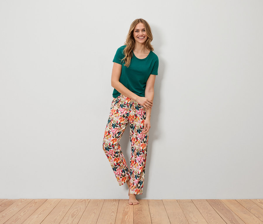 Une femme se tient pieds nus devant un mur. Elle porte un t-shirt vert et un pantalon coloré à motifs de feuilles.