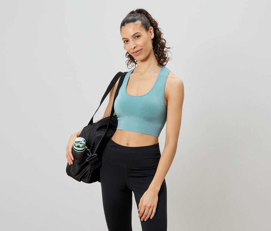 Une femme posant avec un sac de sport, une bouteille d'eau et un soutien-gorge de sport sur un fond clair.
