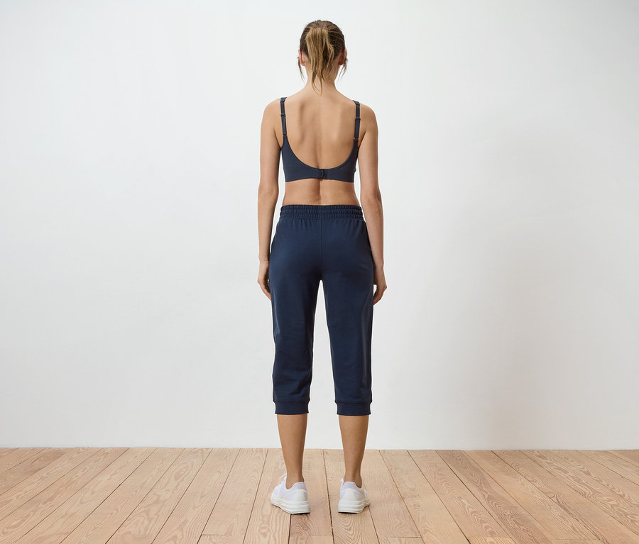 Vue arrière d'une femme aux cheveux blonds attachés portant un soutien-gorge de sport gris et un pantacourt bleu sur un mur blanc.