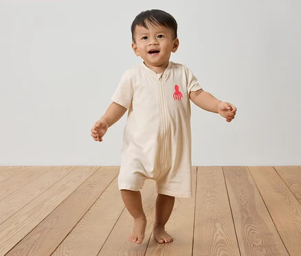 Un jeune enfant marche pieds nus sur un plancher en bois, portant une combinaison beige avec une pieuvre rouge.