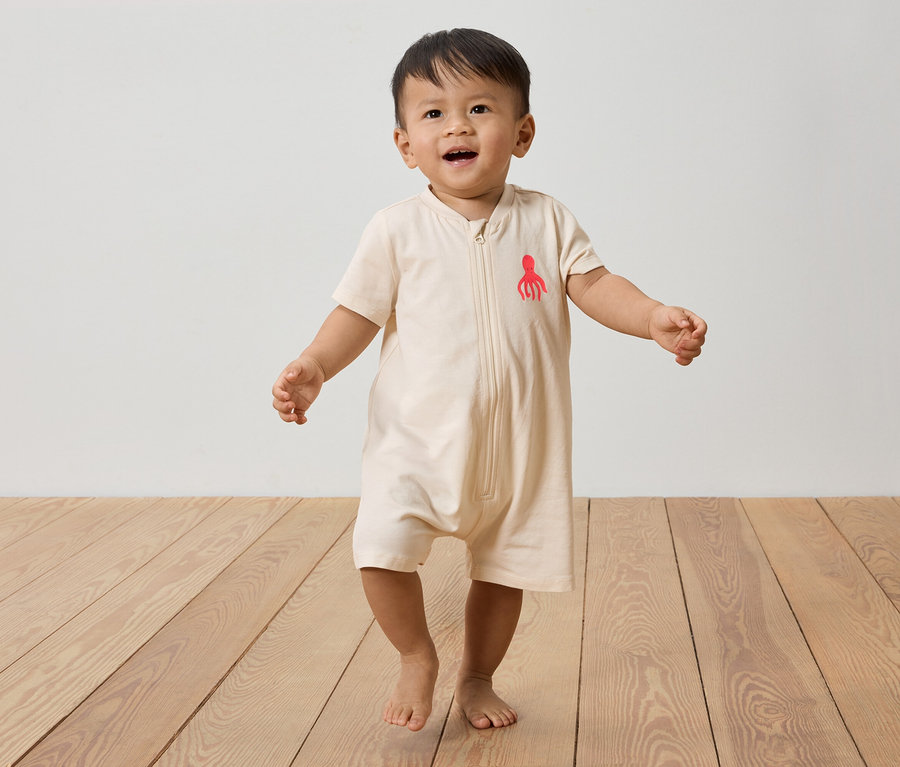 Un jeune enfant marche pieds nus sur un plancher en bois, portant une combinaison beige avec une pieuvre rouge.