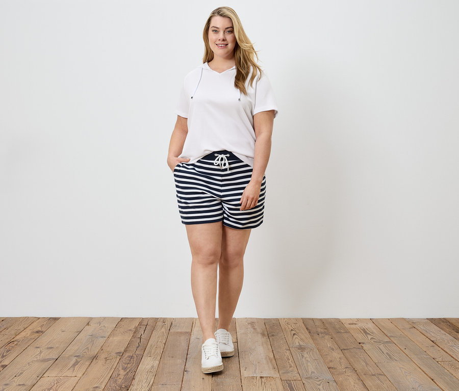 Une femme en t-shirt blanc et short rayé, la main dans la poche, devant un mur blanc et un plancher en bois.