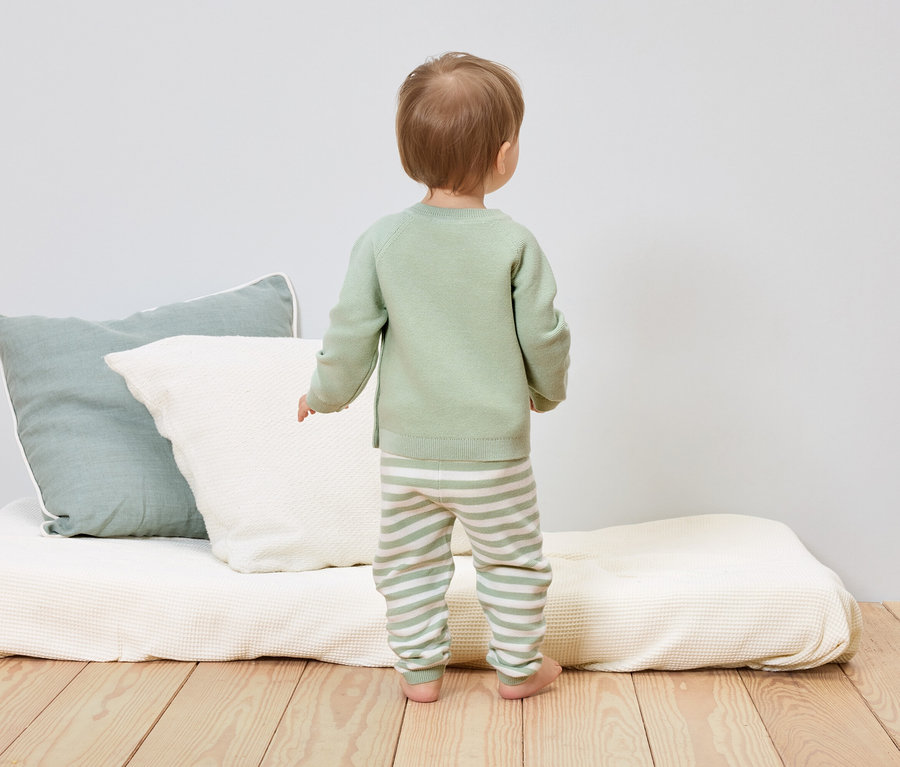 Un enfant en bas âge portant un pull vert clair et un pantalon rayé se tient dos à des coussins et un matelas.