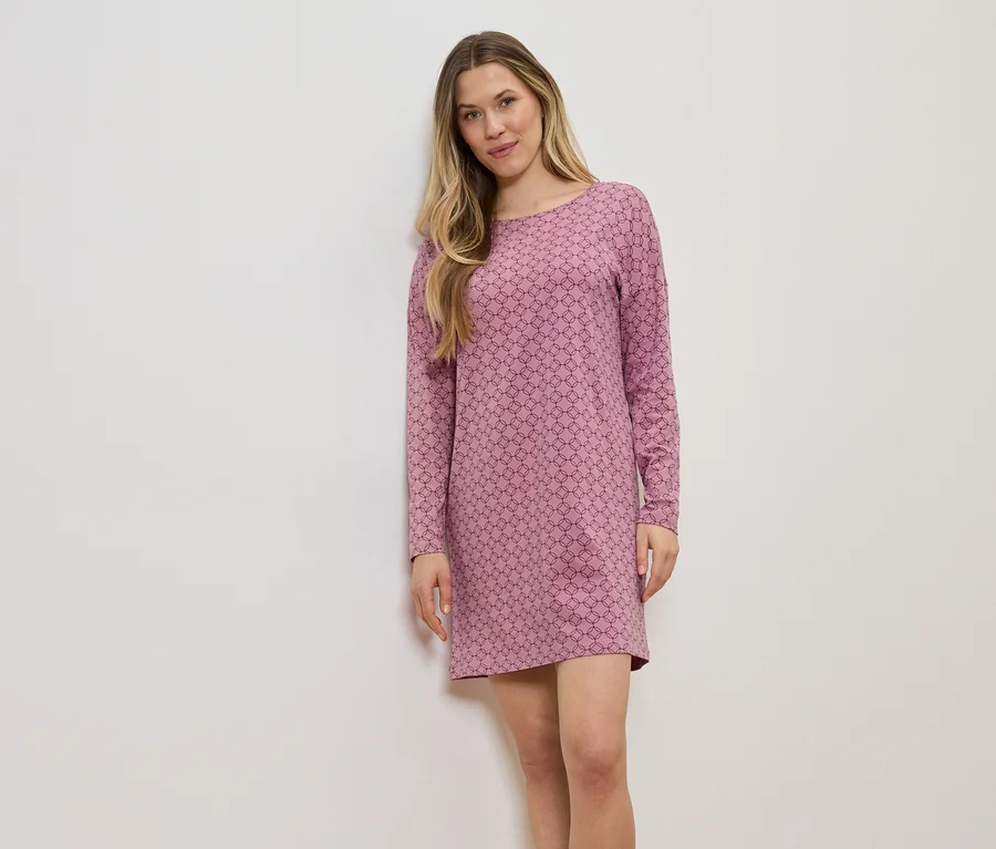 Une femme aux longs cheveux blonds portant une robe rose à manches longues avec un motif géométrique devant un mur blanc.