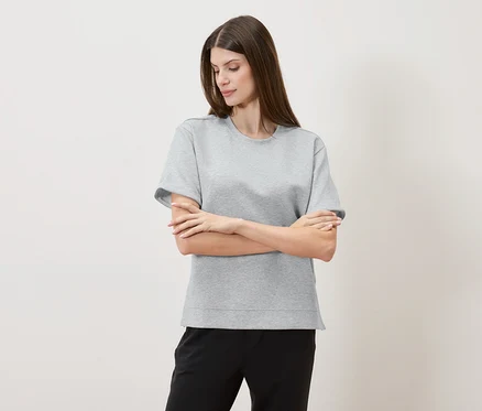 Une femme aux bras croisés porte un t-shirt gris et un pantalon noir devant un fond neutre.