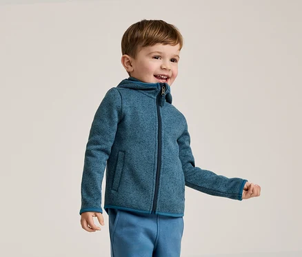 Un garçon souriant aux cheveux bruns porte une veste polaire bleue à capuche et un pantalon de jogging bleu sur un fond clair.