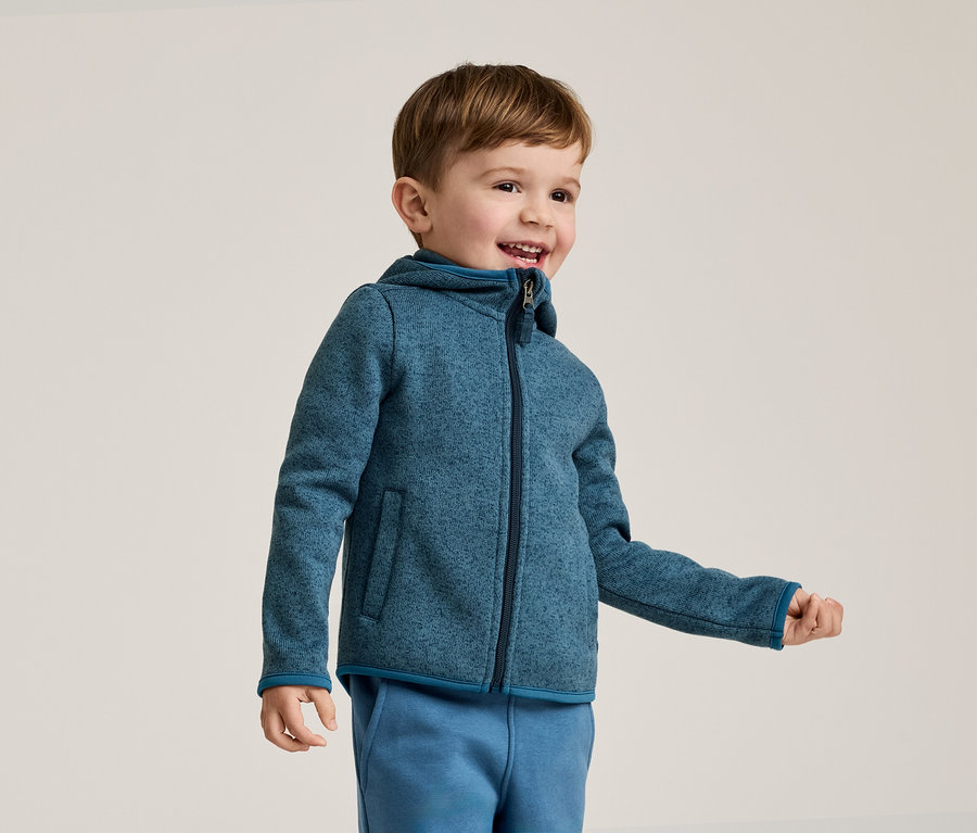 Un garçon souriant aux cheveux bruns porte une veste polaire bleue à capuche et un pantalon de jogging bleu sur un fond clair.