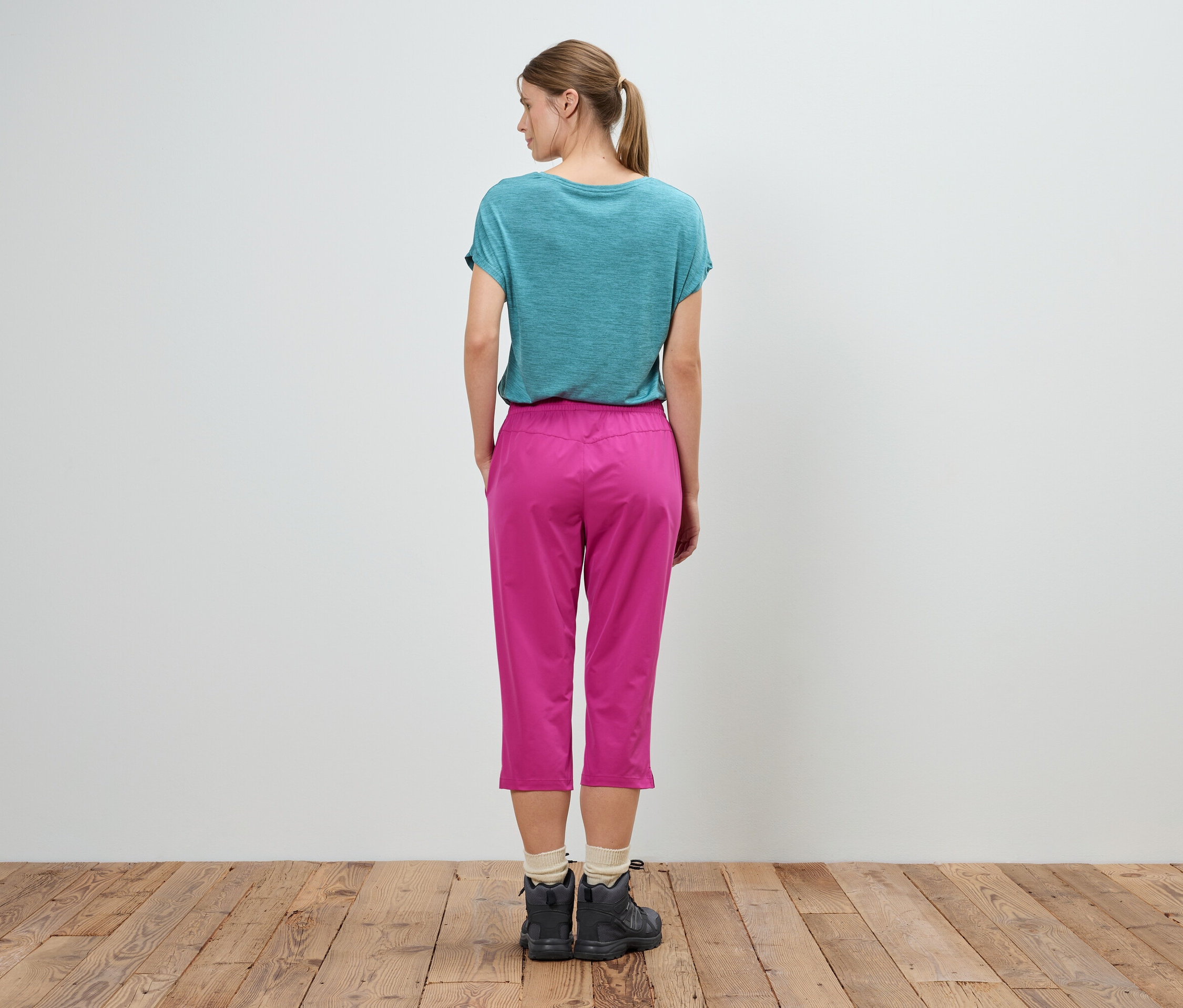 Une femme se tient dos à la caméra, elle porte un t-shirt turquoise, un pantalon rose et des chaussures foncées.
