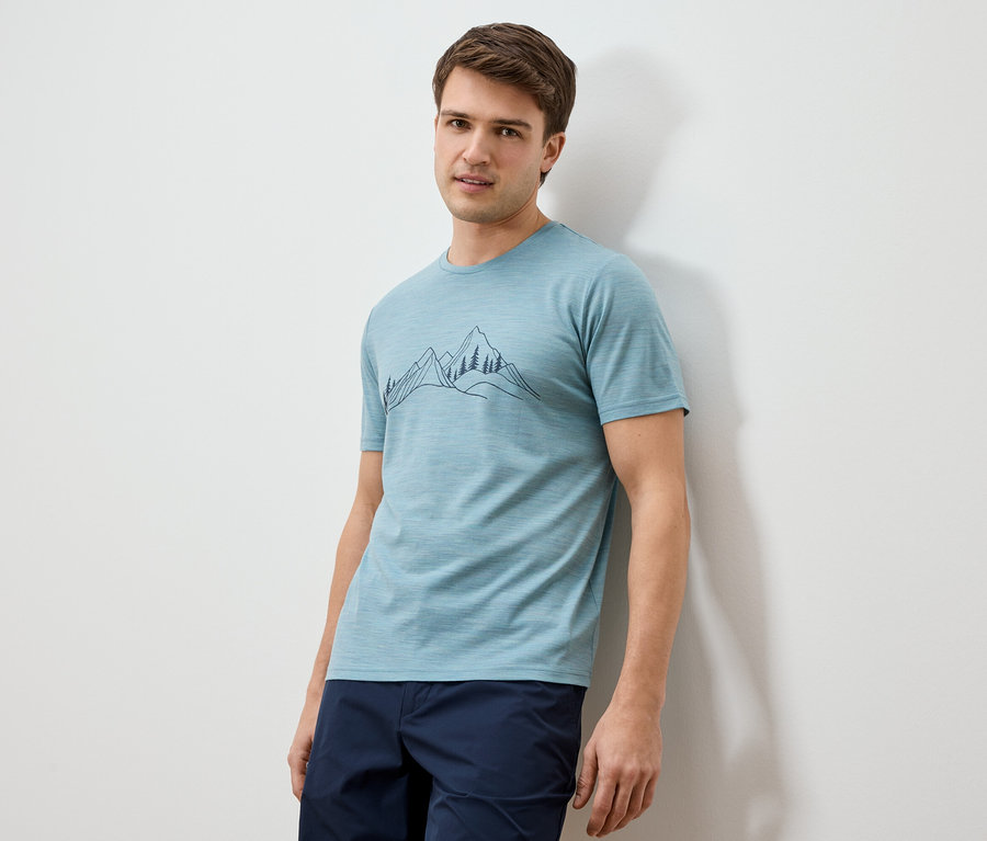 Un homme est appuyé contre un mur et porte un t-shirt bleu clair avec un dessin de paysage de montagne et un short bleu marine.