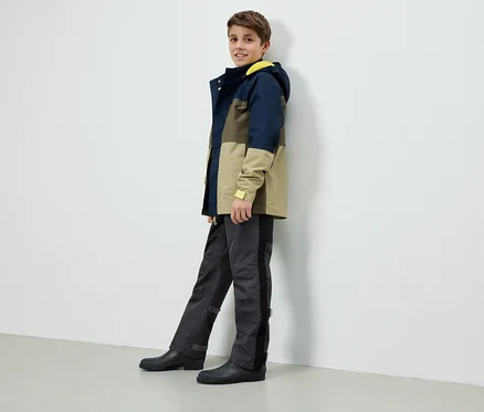 Un garçon se tient contre un mur blanc, portant une veste à blocs de couleurs avec une capuche jaune, un pantalon gris et des bottes en caoutchouc noires.
