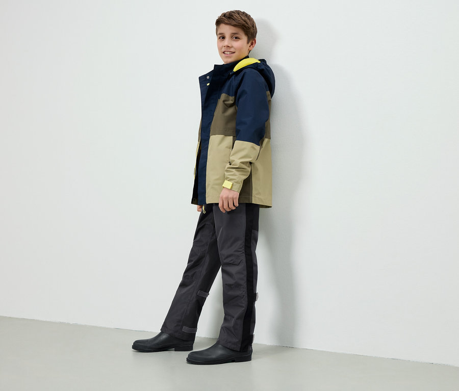 Un garçon se tient contre un mur blanc, portant une veste à blocs de couleurs avec une capuche jaune, un pantalon gris et des bottes en caoutchouc noires.