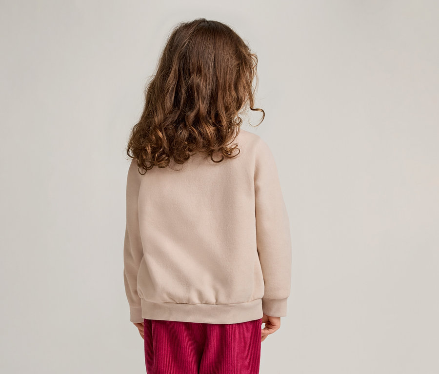 Vue de dos d'un enfant aux cheveux bouclés portant un pull beige et un pantalon en velours côtelé rouge.