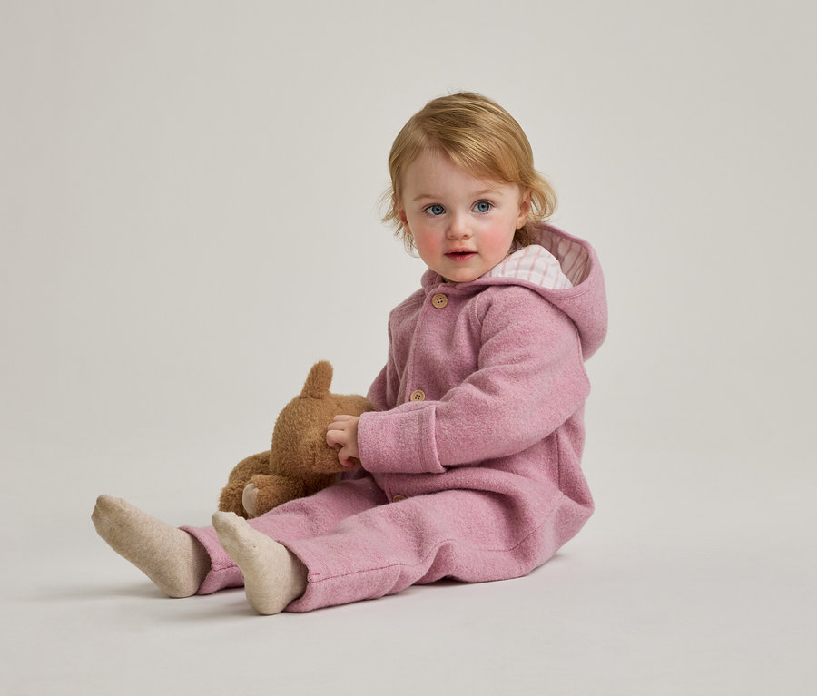 Un bébé blond aux yeux bleus est assis par terre avec un ours en peluche, portant une combinaison rose à capuche.