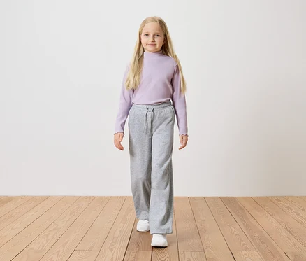 Une fille aux longs cheveux blonds porte un haut à manches longues violet, un pantalon ample gris et des baskets blanches.
