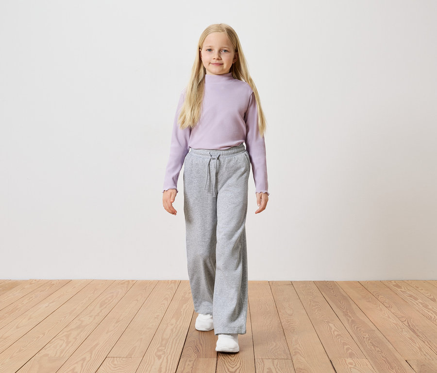 Une fille aux longs cheveux blonds porte un haut à manches longues violet, un pantalon ample gris et des baskets blanches.