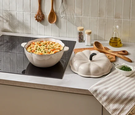 Une casserole crème en forme de citrouille avec des pâtes est sur une cuisinière, à côté se trouve le couvercle. À côté, des herbes, des épices et des ustensiles de cuisine.