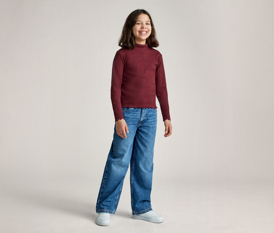 Une jeune fille aux cheveux foncés sourit, vêtue d'un haut bordeaux à manches longues et col montant, d'un jean large bleu et de baskets blanches, posant devant un fond clair.