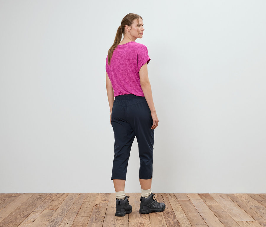 Femme debout dos à la caméra portant un t-shirt rose, un pantalon bleu et des chaussures de randonnée devant un mur blanc.