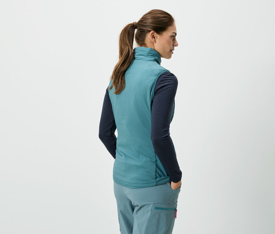 Vue arrière d'une femme avec une queue de cheval portant un gilet turquoise sans manches, une chemise bleue à manches longues et un pantalon turquoise, les mains dans les poches.