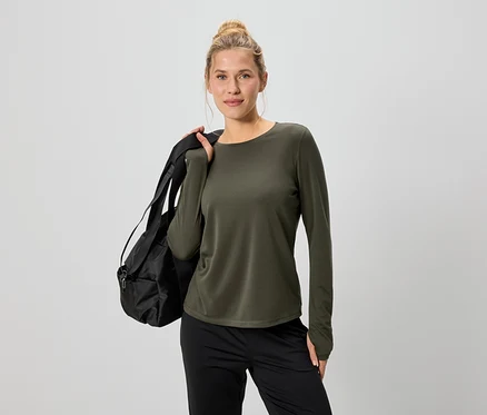 Une femme portant un chignon est vêtue d'un haut à manches longues vert olive et d'un pantalon noir, avec un sac de sport noir sur l'épaule.