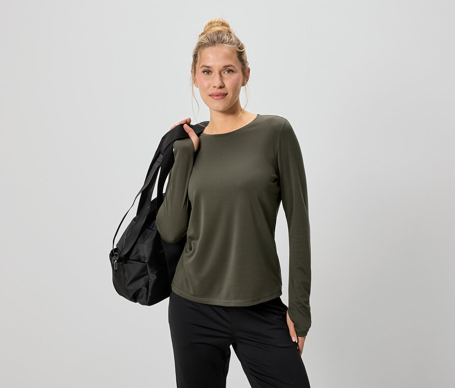 Une femme portant un chignon est vêtue d'un haut à manches longues vert olive et d'un pantalon noir, avec un sac de sport noir sur l'épaule.