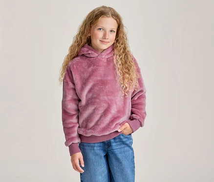 Jeune fille aux longs cheveux blonds bouclés souriante, portant un sweat à capuche rose duveteux et un jean bleu, une main dans la poche.