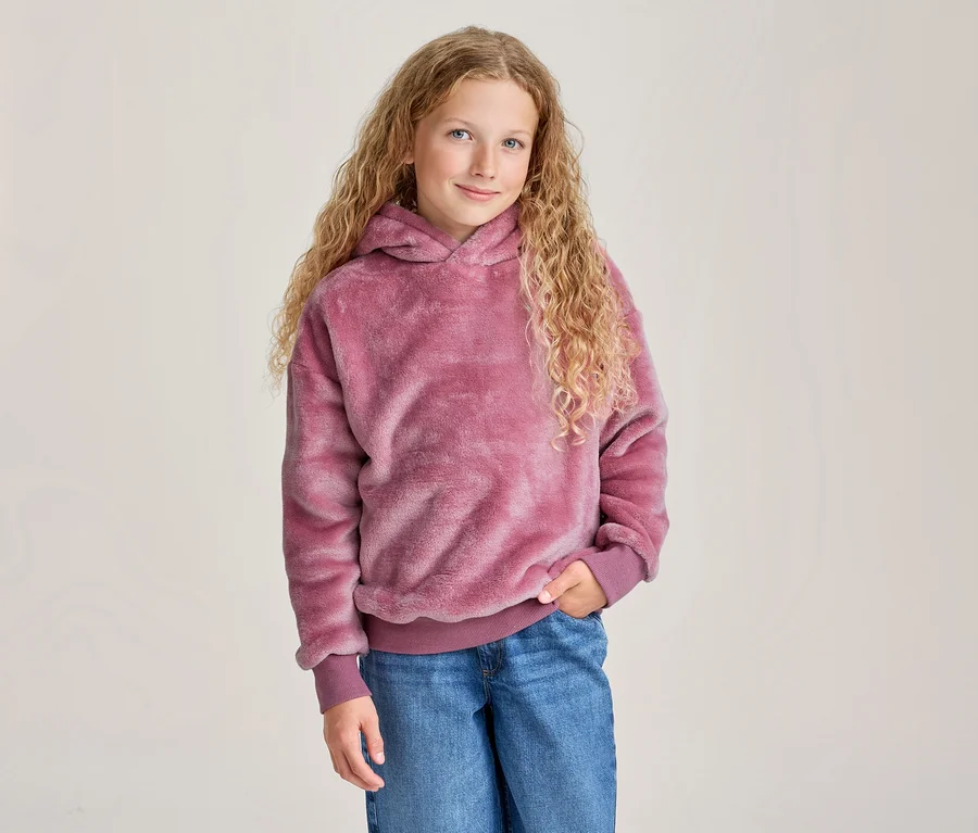 Jeune fille aux longs cheveux blonds bouclés souriante, portant un sweat à capuche rose duveteux et un jean bleu, une main dans la poche.