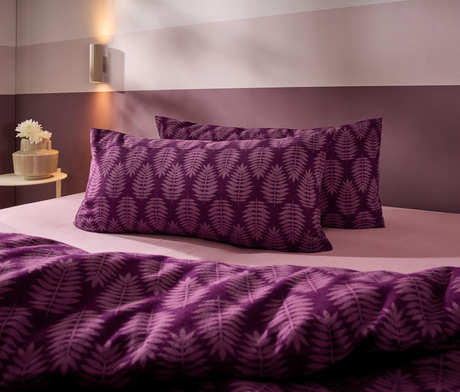 Un lit avec une parure de lit violette à motif de feuilles de fougère, un drap-housse rose clair et un mur violet bicolore. Une applique murale et une table d'appoint avec un vase de fleurs blanches sont visibles.