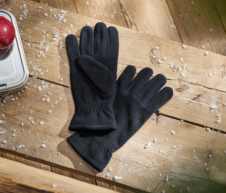 Une paire de gants noirs, une boîte à lunch argentée avec une pomme rouge dessus et des flocons blancs éparpillés sur une surface en bois.