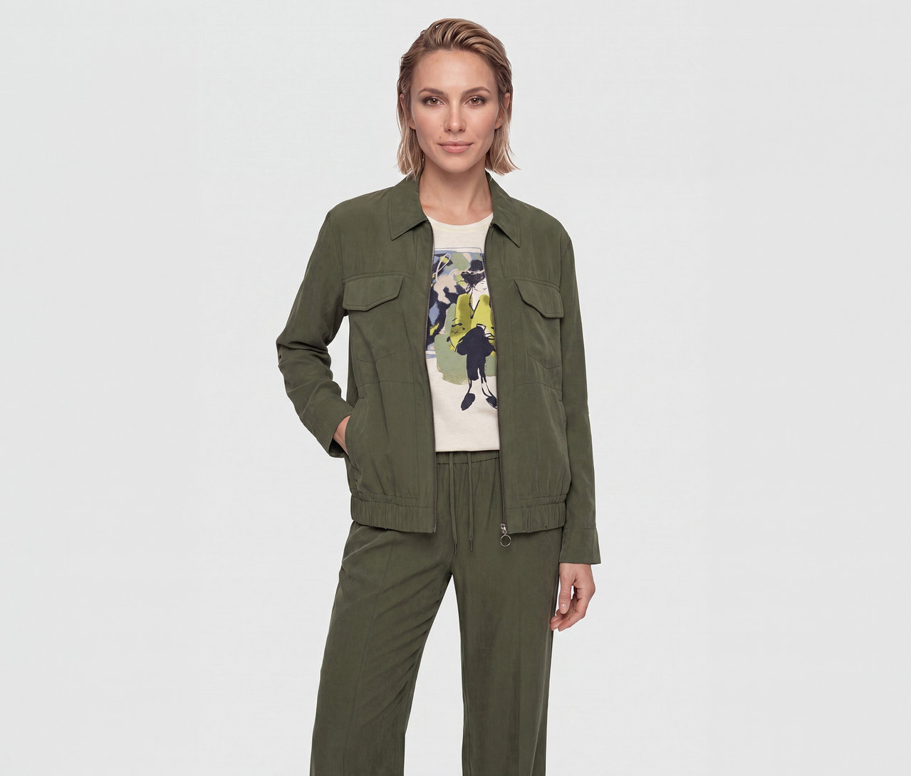 Femme aux cheveux blonds courts portant une veste à col vert olive avec des poches poitrine, un t-shirt crème avec un imprimé graphique coloré et un pantalon vert olive assorti avec cordon de serrage. Sa main gauche est dans la poche de sa veste.