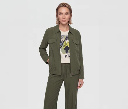 Femme aux cheveux blonds courts portant une veste à col vert olive avec des poches poitrine, un t-shirt crème avec un imprimé graphique coloré et un pantalon vert olive assorti avec cordon de serrage. Sa main gauche est dans la poche de sa veste.