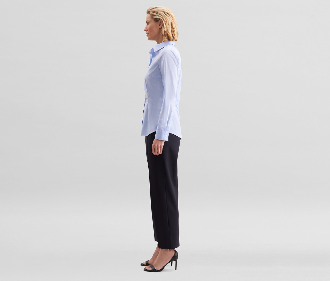 Une femme de profil porte un chemisier Slim Fit non iron Seidensticker bleu clair, un pantalon noir et des sandales noires à talons.