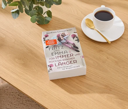 Livre « Fritz und Emma & Für Immer und Noch Ein Bisschen Länger » de Barbara Leciejewski sur table en bois, à côté d'une tasse de café et d'une plante.