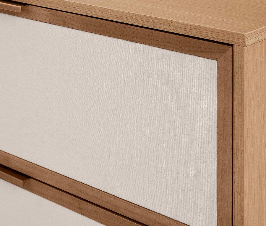 Gros plan d'une commode en bois clair avec deux tiroirs. Les façades des tiroirs sont en tissu crème clair, encadrées de bois, et ont des poignées en bois intégrées.