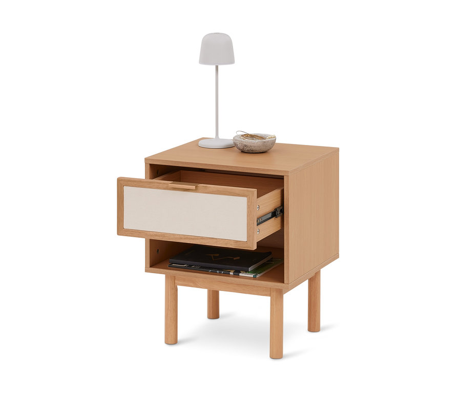 Table de chevet en bois clair avec un tiroir partiellement ouvert à façade crème, une étagère ouverte avec des livres, et une lampe blanche et un bol sur le dessus.