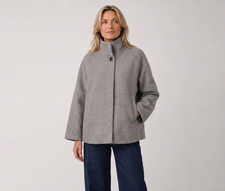Une femme blonde est debout, vêtue d'une veste grise à col montant et manches larges, et d'un jean foncé, la main droite dans la poche.