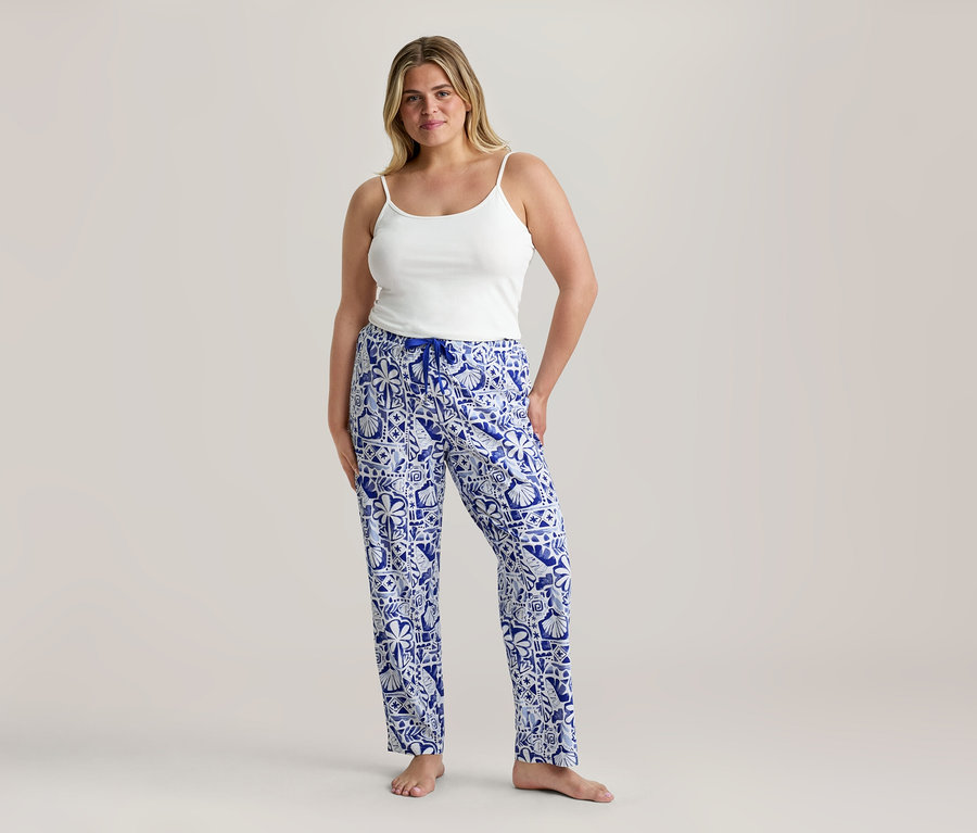 Femme portant un débardeur blanc à fines bretelles et un pantalon de pyjama à motifs bleu et blanc avec un cordon de serrage bleu, debout pieds nus.