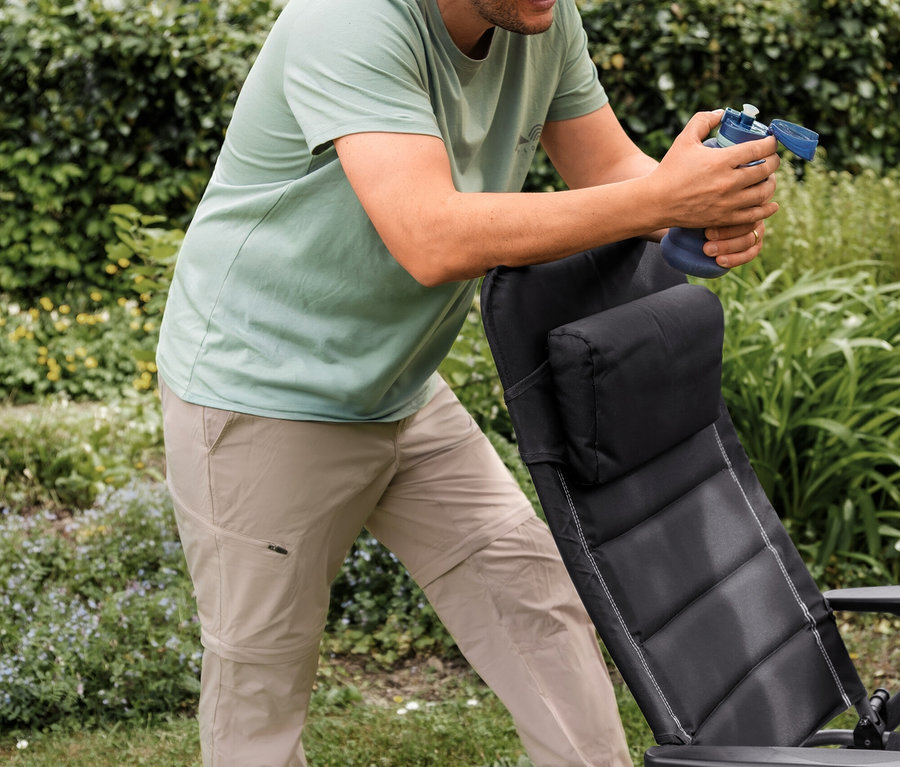 Homme souriant en T-shirt d’extérieur vert menthe et pantalon fonctionnel 2 en 1 beige, appuyé sur une chaise de jardin noire et tenant une bouteille d'eau bleue.