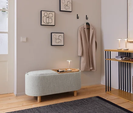 Une entrée accueillante avec un banc rembourré gris clair, sur lequel repose un plateau en bois avec une petite lampe et des clés. Au-dessus, trois tableaux abstraits aux lignes épurées et un manteau beige sont accrochés à des patères noires. À droite, une console en bois avec des supports noirs accueille deux petites lampes, des vêtements pliés, une ceinture et un sac tressé. Un tapis foncé à motifs est posé sur le parquet.