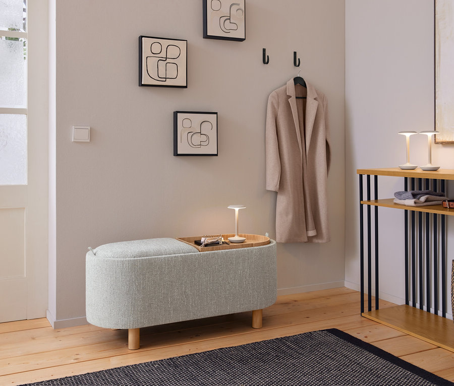Une entrée accueillante avec un banc rembourré gris clair, sur lequel repose un plateau en bois avec une petite lampe et des clés. Au-dessus, trois tableaux abstraits aux lignes épurées et un manteau beige sont accrochés à des patères noires. À droite, une console en bois avec des supports noirs accueille deux petites lampes, des vêtements pliés, une ceinture et un sac tressé. Un tapis foncé à motifs est posé sur le parquet.