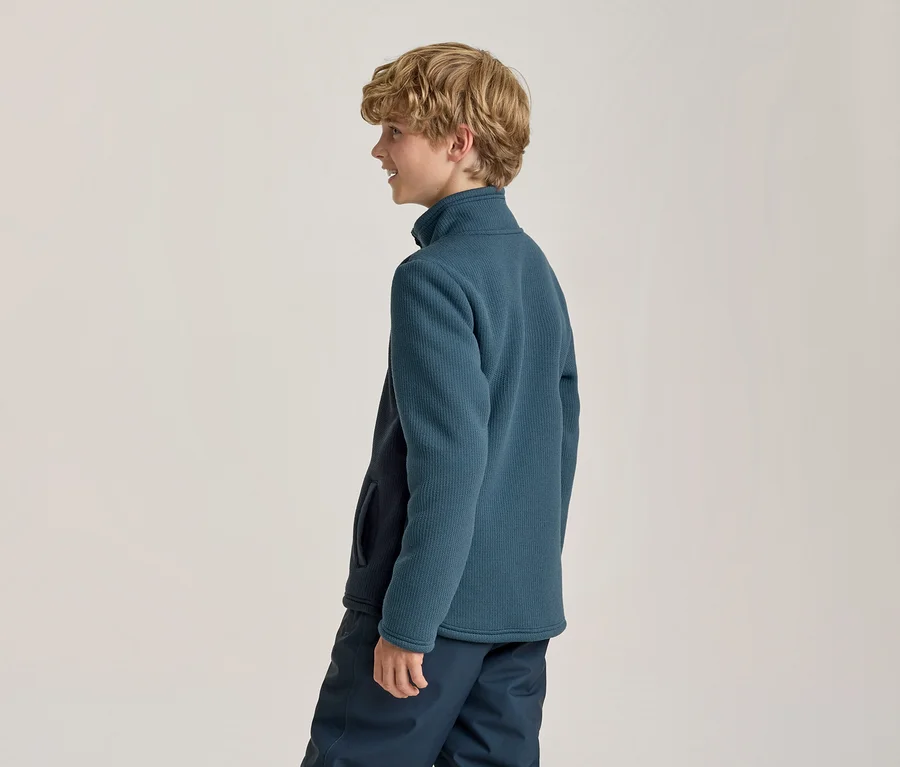 Jeune garçon aux cheveux blonds bouclés, souriant de profil, portant une veste polaire côtelée bleu sarcelle et un pantalon bleu foncé.