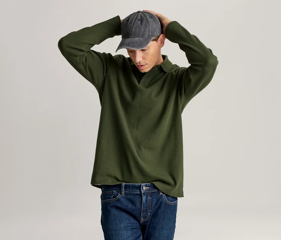 Homme portant un polo vert à manches longues, un jean bleu et une casquette de baseball gris foncé, regardant vers le bas, les mains sur la tête.