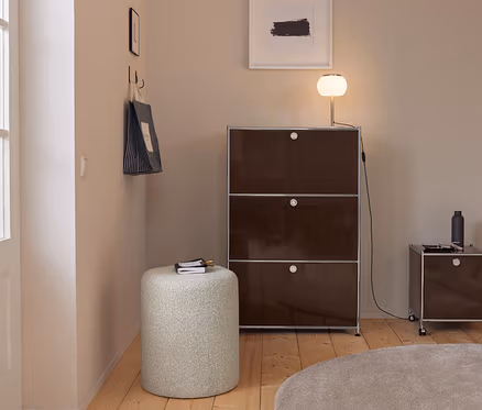 Entrée moderne avec murs clairs, parquet et tapis rond beige. Une grande armoire modulaire marron foncé avec une lampe lumineuse se tient à côté d'une armoire plus petite à roulettes. Un pouf cylindrique clair avec un carnet est au premier plan. Un sac fourre-tout rayé est accroché à des crochets sur le mur gauche.